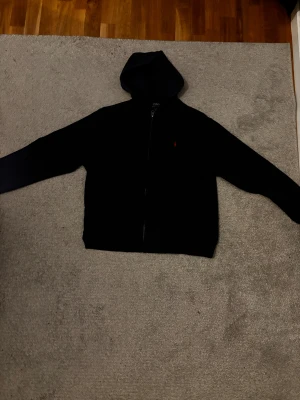 Marinblå hoodie från Polo Ralph Lauren - Marinblå hoodie från Polo Ralph Lauren i storlek 18-20 med klassisk röd broderad logga på bröstet. Tröjan har huva och dragkedja framtill, samt långa ärmar. Perfekt för en avslappnad och stilren look. Pris kan diskuteras vid snabb affär.