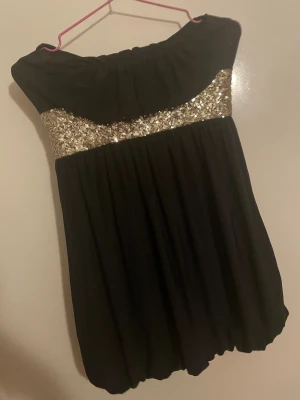 Svart kort klänning med paljettdetalj - Supersnygg svart axelbandslös kortklänning från Bloose med glittrande guldpaljetter över bysten. Klänningen har en luftig, ballongformad nederdel och är perfekt för dig som vill sticka ut. Materialet är mjukt och följsamt, och modellen är loose fit.