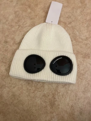 Vit C.P. Company goggle-mössa - Vit ribbstickad mössa från C.P. Company med ikoniska svarta goggle-linser framtill. Klassisk uppvikt kant och cool streetwear-vibe. Tillverkad i mjukt stickat material som håller dig varm och snygg under kalla dagar.