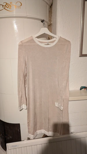 Beige kortklänning med vita detaljer - En stilren beige kortklänning med vita kanter vid halsringning, ärmslut och nederkant. Klänningen har lång ärm och en rak, avslappnad passform. Materialet känns lätt och mjukt mot huden, perfekt för dig som gillar minimalistisk och enkel stil.