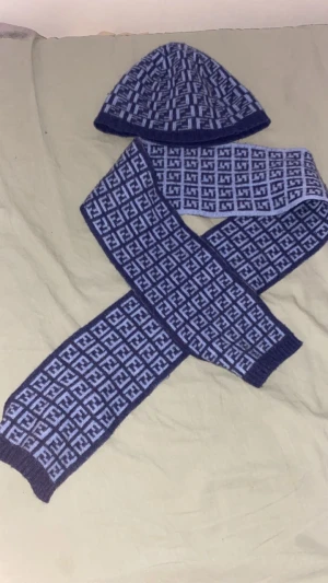 Blå Fendi halsduk med FF-mönster - Snygg halsduk från Fendi i mörkblått och ljusblått med det klassiska FF-logomönstret över hela. Stickad i mjukt material och perfekt för att hålla värmen. Matchande mössa ingår för en komplett look. 