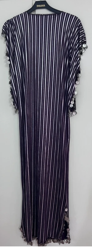 Randig långklänning med myntdetaljer - Långklänning från Indiska med lila och vita ränder, dekorerad med silvriga mynt längs halsringning, ärmar och nederkant. Klänningen har en loose passform och korta ärmar. Materialet känns mjukt och lätt, perfekt för sommarens varma dagar.