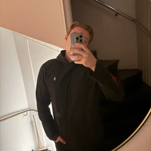 Svart hoodie från Polo Ralph Lauren - Snygg svart hoodie från Polo Ralph Lauren med en ikonisk polospelare broderad på bröstet. Tröjan har långa ärmar och en röd textdetalj på ärmen. Perfekt för en avslappnad stil.