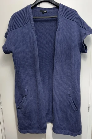 Blå stickad cardigan jacka från Ellos - Snygg blå stickad cardigan jacka från Ellos i storlek 54/56. Jackan har korta ärmar, två fickor fram och en öppen design utan knappar eller dragkedja. Ribbad struktur upptill ger extra stil. Perfekt för lager-på-lager och chill vibe.