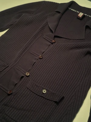 Mörkblå ribbad Pelle P kofta med krage - Snygg mörkblå kofta från Pelle P World Champion med ribbad struktur och klassisk krage. Koftan har fyra bruna knappar framtill, två fickor med lock och knapp, samt ett diskret broderat märke på ärmen. 