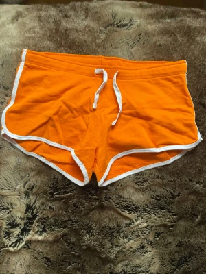 Orange träningsshorts med snörning - Snygga orange shorts med vit kant och snörning i midjan. Modellen är sportig med rundade ben och mjukt bomullsmaterial som är skönt mot huden. Perfekta för träning eller chill. Färgen är riktigt poppig och ger en fräsch vibe.