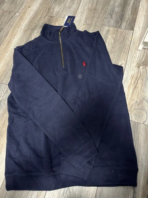 Marinblå half zip tröja Polo Ralph Lauren - Snygg marinblå långärmad tröja från Polo Ralph Lauren med half zip och ståkrage. Tröjan har ett rött broderat Polo-logo på bröstet och är tillverkad i mjuk bomull. Perfekt för dig som gillar klassisk stil med en modern touch.