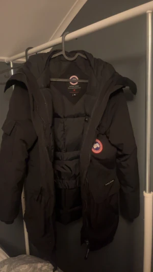 Svart Canada Goose Expedition dunjacka - Canada Goose Expedition dunjacka i svart med klassisk patch på bröstet. Jackan har stor huva, flera fickor och kraftig dragkedja. Quiltad insida och justerbar midja för extra värme. Perfekt för kalla vinterdagar och riktigt snygg streetstil. Päls till luvan finns med. ÄKTA 