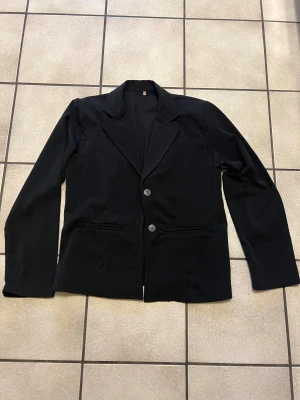 Svart klassisk kavaj med knappar storlek M/L - En svart kavaj med två knappar framtill och klassisk krage. Kavajen har raka ärmar, två fickor framtill och är ofodrad på insidan. Perfekt för dig som gillar en stilren och enkel look.