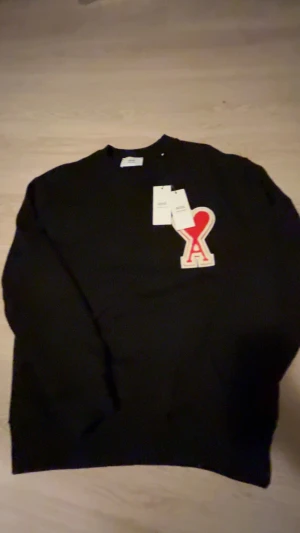 Svart sweatshirt från Ami Paris - Svart sweatshirt från Ami Paris med klassisk rund hals och lång ärm. På bröstet sitter Ami's ikoniska röda hjärta med A-bokstav. Tröjan är tillverkad i mjuk ekologisk bomull och har ribbade muddar vid hals, ärmslut och nederkant. Perfekt för en clean och chill look.