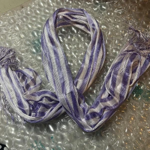 Randig glansig halsduk i silver och lila - Säljer en tunn och glansig halsduk med silvriga och lila ränder. Halsduken har fransar i ändarna och ett lätt skimrande material som ger en cool effekt. Perfekt accessoar för att lyfta din outfit med lite extra färg och shine.