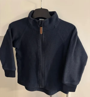 Mörkblå fleecejacka med dragkedja - Mysig mörkblå fleecejacka med hög krage och hel dragkedja framtill. Jackan har långa ärmar och ribbade muddar vid ärmsluten. Perfekt för kyliga dagar när du vill hålla dig varm och bekväm.