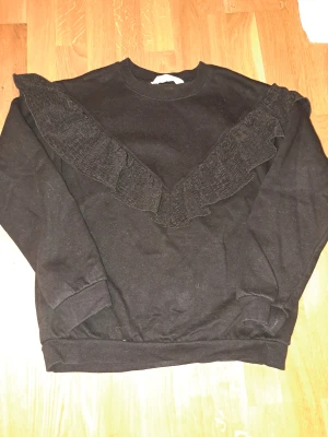 Svart sweatshirt med volang från H&M - Svart sweatshirt från H&M med cool volangdetalj som går i ett V över bröstet och ut på axlarna. Tröjan har rund hals, ribbade muddar och är långärmad. Perfekt för dig som gillar en enkel men ändå unik stil. Både bekväm och vacker!