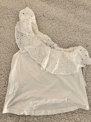 Vit omlott topp med volang från Mango - Söt vit omlott topp från MNG med en stor volang i broderad spets som går över ena axeln. Toppen är axelbandslös på ena sidan och har en ärmlös design. Perfekt för sommarens varma dagar