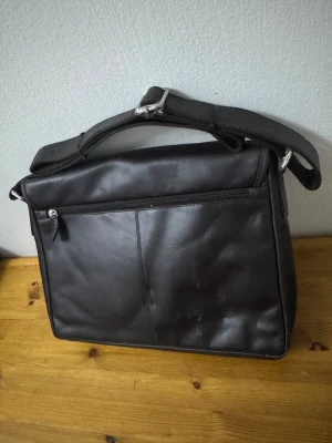 Helt ny och oanvänd Brun datorväska från dbramante1928 - Helt ny och oanvänd Snygg och stilren datorväska från dbramante1928 i mörkbrunt full-grain skinn. Väskan har justerbar axelrem, dragkedja framtill och minimalistisk design. Perfekt för dig som vill ha en klassisk look med plats för laptop och dokument.