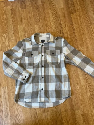Beige och vit rutig skjorta H&M - Snygg rutig skjorta från H&M i beige och vitt med stora rutor. Skjortan har två bröstfickor med knappar, klassisk krage och svarta knappar framtill. Tillverkad i mjuk bomull, perfekt för lager-på-lager. Långärmad och loose fit för chill stil.