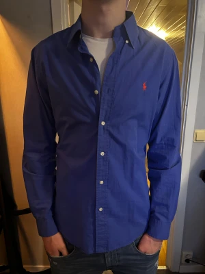 Blå skjorta från Ralph Lauren - Snygg blå skjorta från Ralph Lauren med klassisk krage och vita knappar framtill. Strlk M slimfit, modellen på bild är 193 och väger 82kg. Pris kan diskuteras