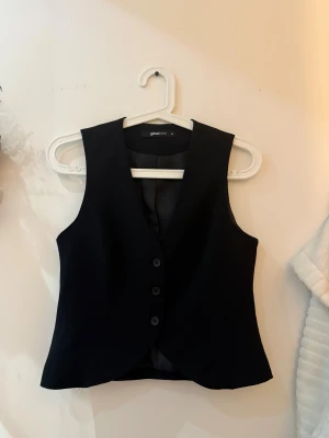 Svart väst från Gina Tricot - Stilren svart väst från Gina Tricot i klassisk modell med tre knappar framtill och v-ringad hals. Västen har en figurnära passform och är tillverkad i ett slätt material som ger en snygg siluett. Perfekt att styla över en skjorta eller t-shirt.