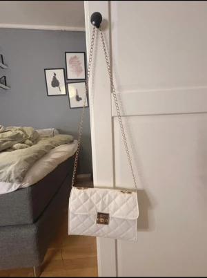 Vit quiltad axelväska med kedja - Snygg vit axelväska med quiltat mönster och guldfärgad kedja. Väskan har ett klassiskt lås framtill och är perfekt för att lyfta din outfit. Insidan är fodrad i beige tyg och väskan har en stilren, rektangulär form.