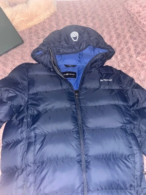 Mörkblå pufferjacka Sail Racing - Säljer en mörkblå pufferjacka från Sail Racing med huva och dragkedja. Jackan har en quiltad design och logga på bröstet. Insidan är fodrad i blått och materialet är syntet. Perfekt för kyliga dagar! Priset kan diskuteras. 