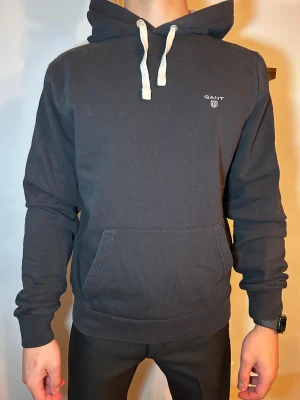 Svart hoodie från GANT med snörning - Svart hoodie från GANT med vit snörning i huvan och liten logga på bröstet. Tröjan har en klassisk känguruficka framtill och är långärmad. Mjuk bomullskänsla och relaxed passform, perfekt för chill dagar.