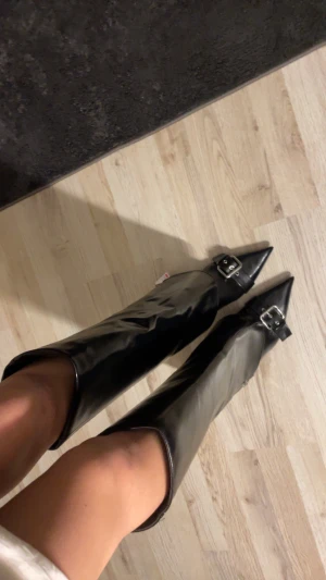 Svarta stövletter med klack från Bershka - Säljer ett par svarta stövletter från Bershka med spetsig tå, hög klack och snygga spänndetaljer framtill. Skorna har dragkedja på sidan och är gjorda i glansigt skinnliknande material. Perfekt för dig som gillar edgy och trendig stil.