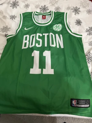 Boston Celtics Irving #11 baskettröja - Grön baskettröja från Nike med vita detaljer, tryckta siffror och text. Framsidan har BOSTON och nummer 11, baksidan IRVING och nummer 11. NBA-logga och GE-märke på bröstet. Klassisk ärmlös modell i lätt polyester, perfekt för sport eller streetwear.