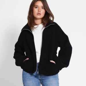 Svart teddyjacka med dragkedja lager 157 - Mysig svart teddyjacka med dragkedja framtill och två stora fickor. Jackan har en avslappnad passform och är perfekt för kyliga dagar. Materialet är fluffigt och ger en cool, casual vibe. Passar dig som gillar streetstyle och vill hålla dig varm.