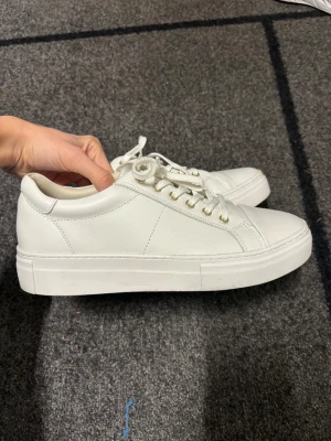Vita sneakers från Vagabond - Fräscha vita sneakers från Vagabond med rund tå och platt sula. Skorna har snörning med vita skosnören och guldfärgade öljetter. Tillverkade i slätt skinn för en clean look. Perfekta för dig som gillar minimalistisk stil och vill ha ett par klassiska sneakers.