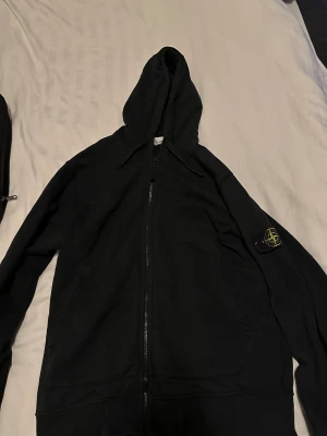 Svart hoodie från Stone Island - Svart hoodie från Stone Island med dragkedja och huva. Klassisk patch med logga på vänster ärm. Tröjan har två fickor framtill och snörning i huvan. Perfekt för dig som gillar streetwear och vill ha en clean look.