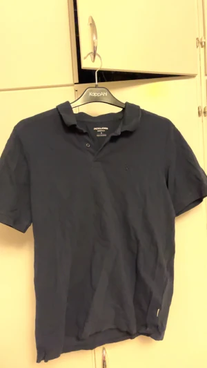 Marinblå pikétröja Jack & Jones S - Snygg marinblå pikétröja från Jack & Jones Essentials i storlek S. Klassisk krage med två knappar framtill och korta ärmar. Tillverkad i mjuk bomull som är skön mot huden. Perfekt för dig som gillar stilrena och enkla plagg.