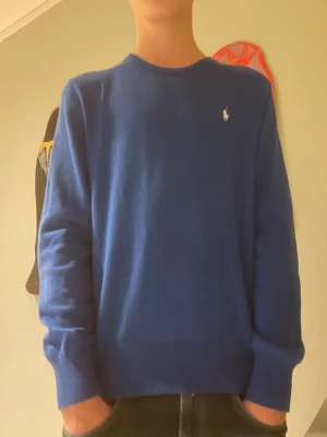 Blå stickad tröja från Polo Ralph Lauren, jr L men passar XS - Storken L 14-16 men passar XS. Klassisk blå stickad tröja från Polo Ralph Lauren med rund halsringning och broderad vit logga på bröstet. Tröjan har ribbade muddar vid ärmslut och nederkant, perfekt för en stilren och enkel look. Knappt använd och är i väldigt fint skick. Pris kan diskuteras, skriv vid funderingar