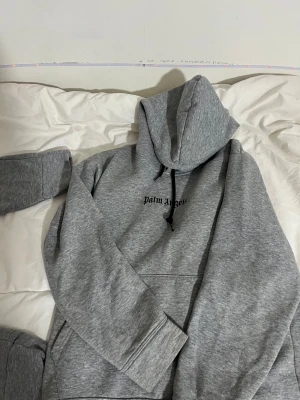 Grå Palm Angels hoodie med tryck - Den är använd några gånger.      Säljer en grå hoodie från Palm Angels med svart logga på bröstet och stort grafiskt tryck på ryggen. Tröjan har huva med svarta snören, lång ärm och en klassisk känguruficka framtill. Perfekt för dig som gillar streetwear och snygga detaljer.