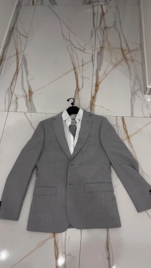 Grå kostym slim fit från Dressmann - Snygg grå kostym från Dressmann i slim fit-modell. Kavajen har klassisk krage, två knappar och fickor med lock. Byxorna är raka och matchar kavajen perfekt. Materialet är mjukt och lätt, perfekt för dig som vill ha en stilren look. Passar dig som gillar enkel och modern stil. Kostymen har varit använd 2 gånger till skolan 