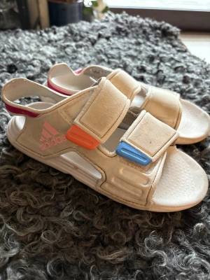 Sandaler från Adidas med färgdetaljer - Säljer ett par sandaler från Adidas med coola färgdetaljer i orange, blått och rosa. Sandalerna har två breda kardborreband och öppen tå, perfekt för varma dagar utomhus eller kalla vinterdagar inomhus. Adidas-loggan syns på sidan och materialet är syntet för lätt och bekväm känsla.