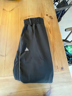 Svarta täckbyxor baggy - Svarta täckbyxor med resår i midjan och benslut, samt två knappar framtill. Helt nya har aldrig använt. Byxorna har en vit logga på vänster ben och diskret piping längs sidan. Perfekta för kalla dagar och utomhusaktiviteter.