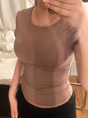 Brun tight kortärmad topp - Säljer en brun, tight och figurnära topp med korta ärmar och rund halsringning. Toppen har en slät yta och är croppad i modellen, vilket ger en snygg siluett. Perfekt för dig som gillar enkla och stilrena plagg med modern passform.