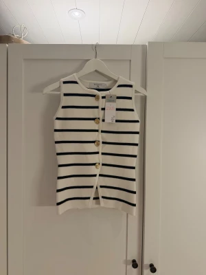 Vit och marinblå randig linnetopp Primark - Säljer en fräsch ärmlös linnetopp från Primark i storlek 2XS men passar mig som brukar ha XS/S. Toppen är vit med marinblå horisontella ränder och har fyra snygga guldfärgade knappar framtill. Materialet är mjukt och stickat, perfekt för en chill och stilren look.