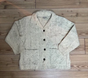 Beige stickad cardigan jacka L - Säljer en beige stickad cardigan jacka från Weave of the Irish, handgjord på Irland. Jackan har grovstickat mönster med mörka knappar och två stora fickor framtill. Materialet är 100% akryl och den har en klassisk krage. Perfekt för dig som gillar chill och mysig stil.