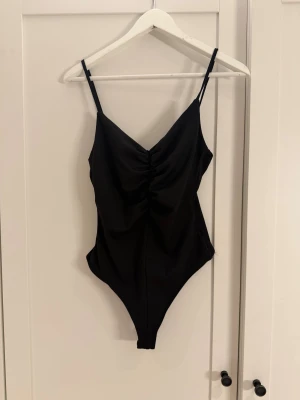 Svart body med rynkad front - Snygg svart body med smala axelband och rynkad detalj framtill som ger en cool och modern look. Modellen har en v-ringad form och är tillverkad i mjukt syntetmaterial som sitter skönt mot kroppen.