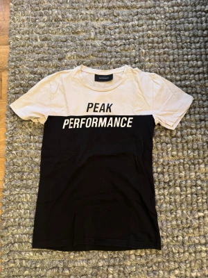 Peak Performance svartvit t-shirt - Snygg t-shirt från Peak Performance i svart och vitt med stor logga över bröstet. Klassisk rund halsringning och korta ärmar. Tillverkad i mjuk bomull för en skön känsla. Perfekt för en sportig och stilren look.