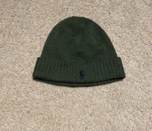 Grön stickad mössa från Polo Ralph Lauren - Snygg mörkgrön mössa från Polo Ralph Lauren i mjuk och tvättbar merinoull. Klassisk ribbstickad design med uppvikt kant och broderad Polo-logga framtill. Perfekt för dig som vill ha en stilren och varm accessoar till hösten och vintern.