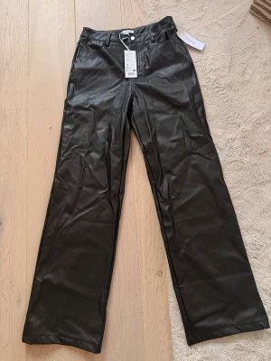 Svarta wide byxor i skinnimitation 36  - Snygga svarta byxor från Bubbleroom i skinnimitation med hög midja och raka, vida ben. Klassisk femficksmodell med bälteshällor och knappstängning framtill. Perfekt för dig som vill ha en edgy och trendig look.