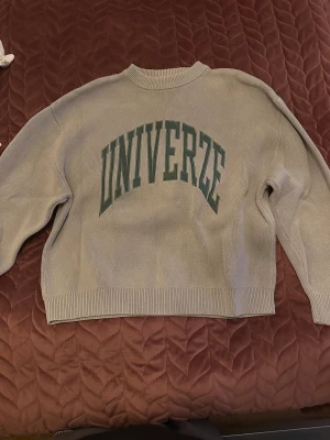 Beige stickad tröja WEEKDAY M - Snygg beige stickad tröja från WEEKDAY i storlek M. Tröjan har grönt tryck med texten 'UNIVERZE' på framsidan, ribbad hals och muddar samt långärmad passform. Perfekt för dig som gillar clean och enkel stil med en twist.