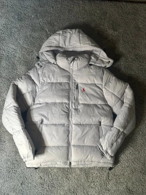 Ljusgrå pufferjacka från Polo Ralph Lauren - Säljer en ljusgrå pufferjacka från Polo Ralph Lauren med huva och broderad röd logga på bröstet. Jackan har dragkedja framtill, två sidofickor med dragkedja och quiltad design. Perfekt för kalla dagar och har en clean, modern look.
