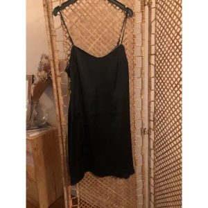 Svart satinklänning NA-KD x Moa Mattsson - Svart kortklänning i satin från NA-KD x Moa Mattsson med tunna axelband och glittriga stenar längs halsringningen. Enkel och stilren design som känns lyxig och trendig. Perfekt för dig som gillar att sticka ut med detaljer. Liten i storlek.