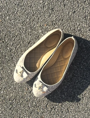 Beige ballerinaskor Michael Kors - Snygga ballerinaskor från Michael Kors i beige med diskret MK-mönster. Skorna har rund tå, quiltad innersula och en dekorativ rosett med guldfärgade detaljer framtill. Perfekta för dig som gillar stilrena och trendiga accessoarer.