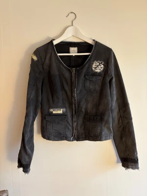 Svart jeansjacka från Cream - Cool svart jeansjacka från Cream med flera broderade patches och detaljer i silver. Jackan har dragkedja framtill, råa kanter och fickor med lock. På ena ärmen finns svarta och silvriga pärlor. Perfekt för dig som gillar en edgy stil.