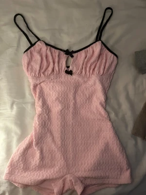 Rosa ärmlös korsett med rosetter - Supersöt rosa korsett med tunna svarta axelband och två små svarta rosetter framtill. Materialet är mjukt och har en strukturerad yta som ger en unik look. Toppen är ärmlös och har en feminin form med svart kant runt bysten.