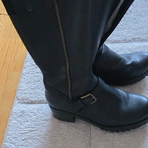 Svarta höga boots med spänne, strl 39 - Säljer ett par svarta boots med grov sula och låg klack. De har dragkedja längs sidan och ett snyggt spänne över foten. Utsidan är i skinn och insidan är fodrad med mjukt material. Perfekta för höst och vinter när du vill ha både stil och komfort.